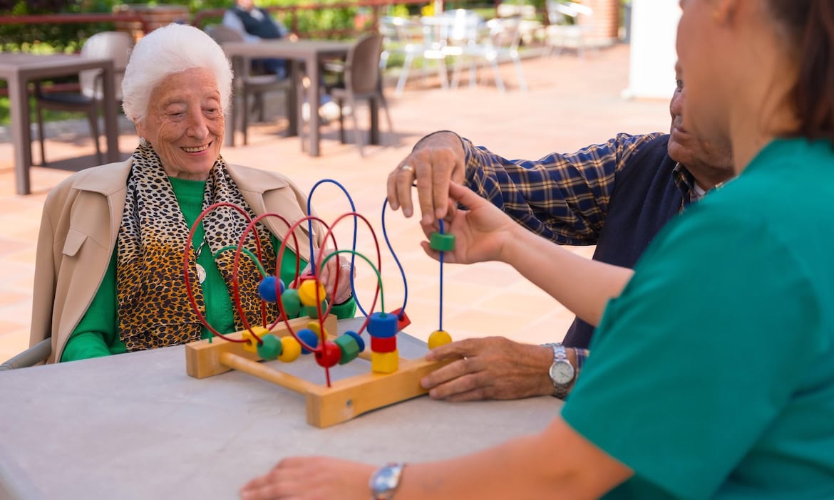 Pflegerin spielt mit zwei älteren Menschen: EIne Pflegeversicherung übernimmt einen Teil der Kosten für einen Aufenthalt im Pflegeheim.