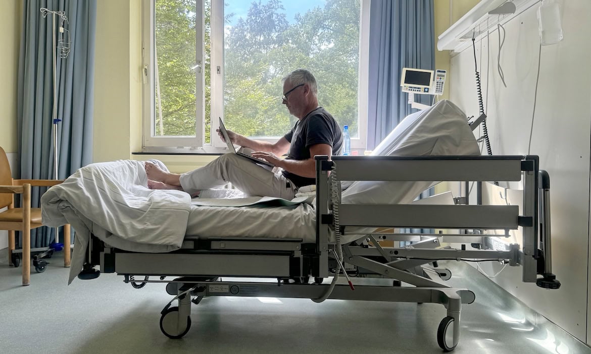Mann am Laptop im Einzelzimmer im Krankenhaus: Die Beiträge vieler PKV-Versicherter steigen 2026 zweistellig.